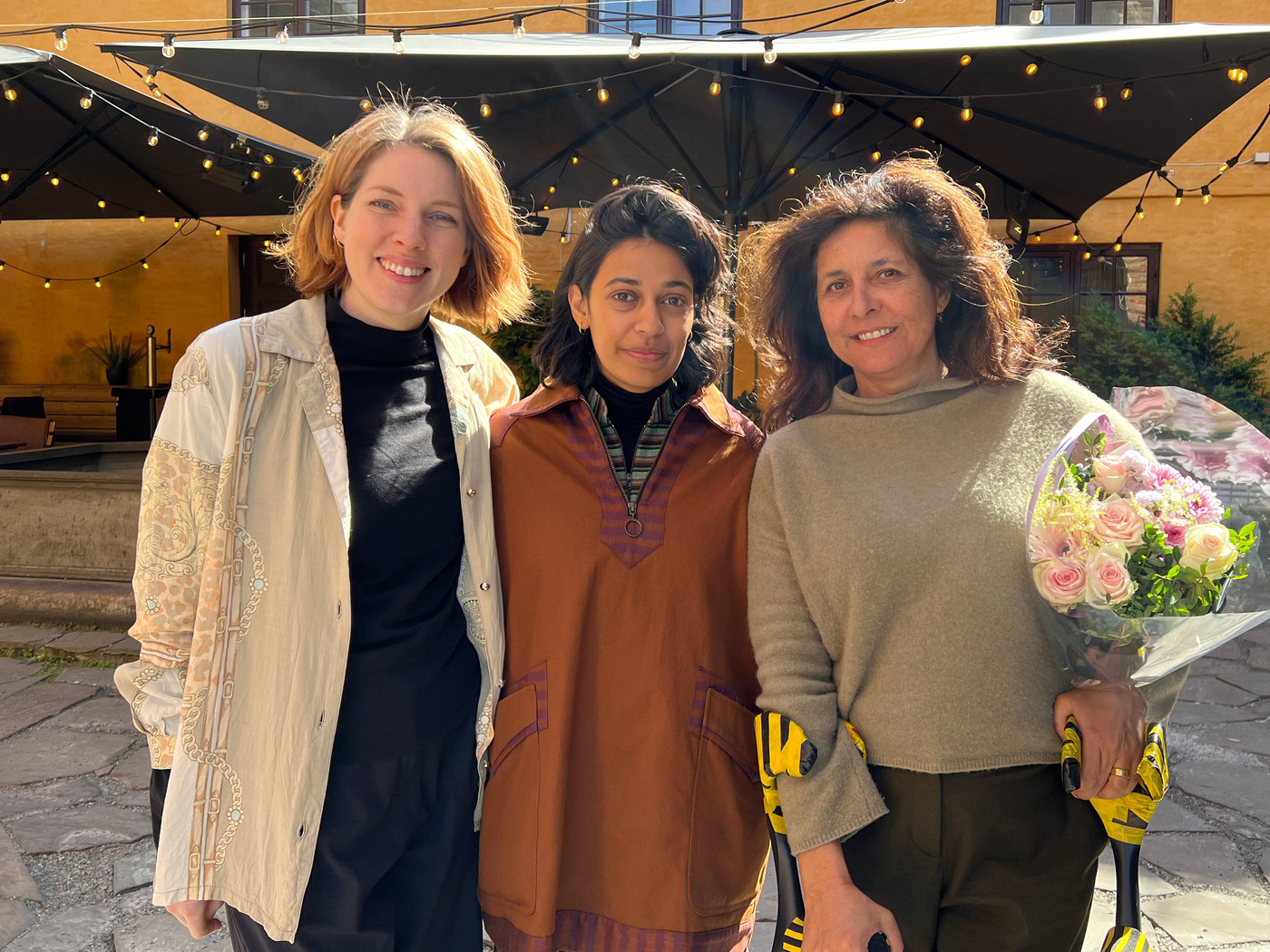 Tre kvinner smiler og poserer utendørs foran en gul bygning. Kvinnen til venstre har lyst, skulderlangt hår og er iført en lys, mønstret jakke over en svart genser og et langt, mørkt skjørt. Kvinnen i midten har mørkt, skulderlangt hår og er iført en brun tunika med broderier over en stripet genser. Kvinnen til høyre har mørkt, krøllete hår og holder en stor bukett med lyse roser innpakket i plast. Hun er iført en grå genser og mørke bukser. Bak dem er det en gul bygning med flere vinduer og svarte markiser med lyslenker under. Bakken er dekket av lyse steinheller.