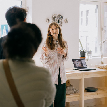 En ung kvinne tar imot to gjester i atelieret. I bakgrunnen en laptop og skulpturer.