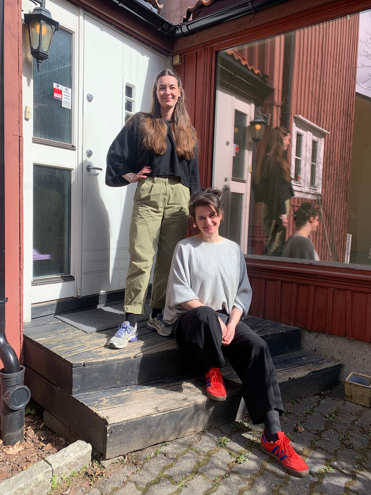 To kvinner på en solfylte trapp foran en rød trebygning. Sophie står på øverste trinn, og Jasmina sitter foran henne.