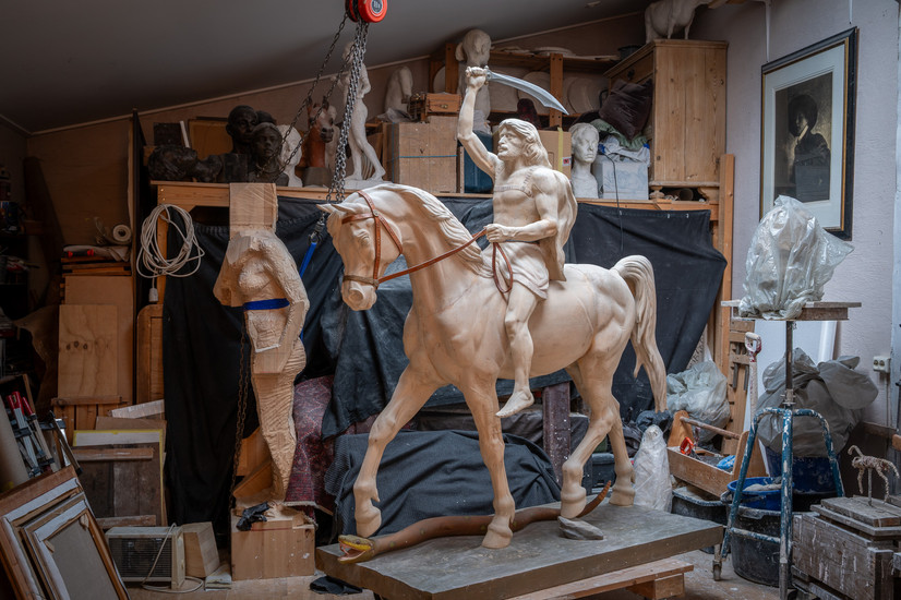 Utvidet bildebeskrivelse (for synshemmede):

Bildet viser et stort figurativt skulpturarbeid inne i et kunstneratelier. I sentrum står en monumental modell av en mann til hest, utført i et lyst materiale som leire eller gips. Hesten er fremstilt i bevegelse, med løftet forbein og spent kropp, mens rytteren sitter barføtt på ryggen. Mannen har langt hår og skjegg, og holder en krummet sabel hevet over hodet i høyre hånd, som i et dramatisk øyeblikk av kamp eller triumf. Den venstre hånden holder tøylene. Kroppen er muskuløs og anatomisk detaljert, med tydelig markerte armer, bryst og ben.

Skulpturen står på en solid arbeidsplattform i tre. Rundt verket er atelieret tett fylt av arbeidsmaterialer og andre skulpturer. I bakgrunnen ses flere byster og figurer, noen ferdige og andre under arbeid, plassert på hyller og benker. Til venstre står en uferdig, grovt tilhugget figur i tre, delvis surret med tape. Over skulpturen henger en kjetting og en talje, brukt til løfting av tunge arbeider. Vegger og hyller er fylt med verktøy, former, plastdekkede gjenstander og støpematerialer. En innrammet grafikk henger på veggen til høyre.

Rommet har dempet, naturlig lys og gir et sterkt inntrykk av et aktivt arbeidssted. Helheten formidler kunstnerisk prosess, håndverk og konsentrasjon, med fokus på klassisk figurativ skulptur, anatomi og bevegelse.

SEO-optimalisert bildebeskrivelse:

Figurativ rytterstatue under arbeid i kunstneratelier. Stor skulptur av mann til hest med hevet sabel, modellert i leire eller gips, omgitt av byster, verktøy og arbeidsmaterialer. Klassisk realistisk billedhuggerkunst med fokus på anatomi, bevegelse og historisk motiv, fotografert i skulptørens atelier.