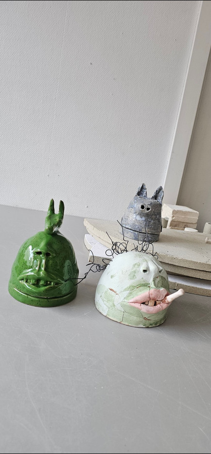• Bildet viser håndlagde keramikkskulpturer.

• En grønn klokkeformet figur har et uttrykksfullt ansikt og hornlignende utvekster.

• En hvit/grønnaktig figur er et fragmentert hode med lepper og en pinne i munnen, holdt sammen med metalltråd.

• Bak er en mindre, gråaktig figur som ligner et stilisert katte- eller hundeansikt med spisse ører og øyehull.