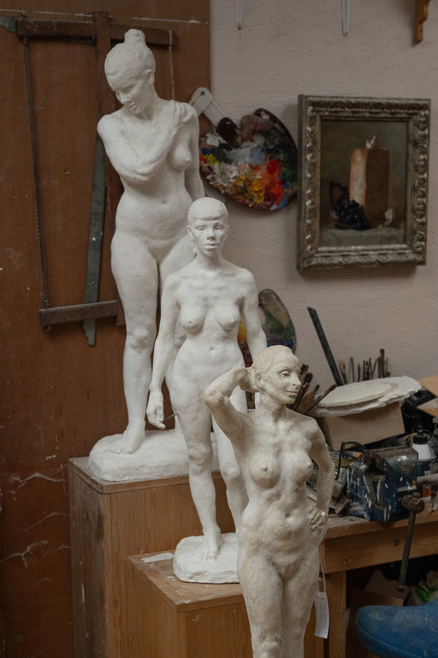 Utvidet bildebeskrivelse (for synshemmede):

Bildet viser tre figurative skulpturer av nakne kvinneskikkelser plassert inne i et kunstneratelier. Skulpturene er modellert i et lyst materiale, som gips eller leire, og varierer i størrelse og positur. To stående kvinneskikkelser er plassert på trepidestaller i bakgrunnen. Begge figurene er fremstilt stående i rolige, innadvendte positurer med tydelig anatomisk bearbeiding. Den ene har armene krysset foran brystet og hodet svakt bøyd nedover, mens den andre står med armene naturlig ned langs siden og blikket rettet fremover med et nøytralt, rolig uttrykk.

I forgrunnen står en mindre kvinneskikkelse, plassert noe foran de øvrige. Hun holder den ene hånden bak hodet og den andre i siden, noe som skaper en klassisk kontrapoststilling som fremhever kroppens linjer og bevegelser. Uttrykket hennes oppleves som avspent og selvsikkert.

Rundt skulpturene sees elementer fra kunstnerens arbeidsmiljø. På veggen bak henger en malerpalett med tørkede farger i mange nyanser. Til høyre er et innrammet stillebenmaleri med frukt og et stearinlys. Under dette står en arbeidsbenk med pensler, modelleringsverktøy, beholdere og materialer, som gir et tydelig inntrykk av et aktivt og levende atelier. Lyset er dempet og mykt, og fremhever overflater, teksturer og detaljer i skulpturene, og bidrar til en stille, konsentrert og kontemplativ stemning.

SEO-optimalisert bildebeskrivelse:

Figurative skulpturer av nakne kvinneskikkelser i gips eller leire plassert i kunstneratelier. Tre stående kvinnefigurer utført i klassisk realistisk stil med fokus på anatomi, proporsjoner og kontrapost. Skulpturene er omgitt av kunstnerverktøy, paletter og atelierutstyr, og viser den skapende prosessen innen tradisjonell figurativ billedhuggerkunst.