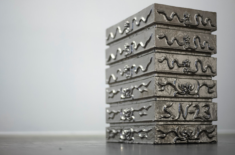 Glazed stoneware sculpture coated with platinum luster. The sculpture is rectangular and composed of multiple rectangles that appear to be stacked on top of each other. Each rectangle features ornaments, with a flower-like formation at the center of each ornament. The top of the sculpture consists of a surface with a hole. Through this hole, you can see down into yet another hole.