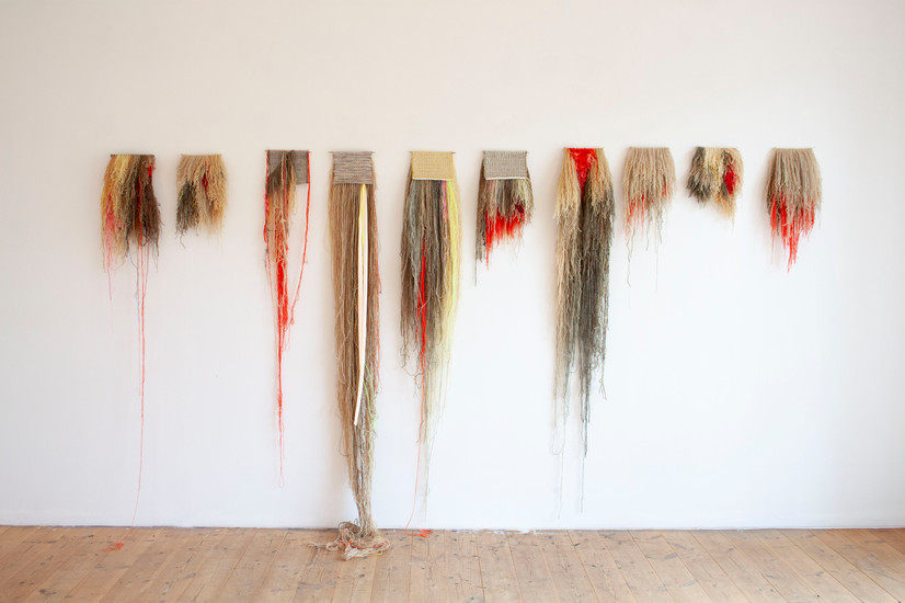 Ti småe rye tepper på rad, fargene er rød, beige, gul, brun og grønn. Første teppe er kort med noen få lange røde tråder, neste er kort, deretter noen lange røde tråder, to neste har en veldig lang lugg som går helt ned til gulvet og ryene er snudd slik at baksidens flatvev er fram, neste er kort, men også snudd slik. Da er det en lang igjen, men luggside er synlig. Tre siste er korte med luggsiden fremst.  