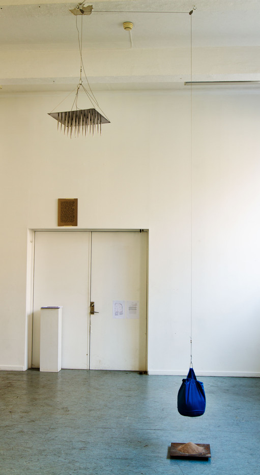 På bilden syns en metallplate av skarpe pigger hengende over hodene til publikum, holdt oppe av ståltau koblet til et motvekt av sand som sakte lekker ut fra posen sin