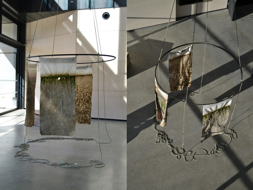 Tre tekstiler som henger rundt en stor metallring i et åpent rom. Installasjonen er stor nok til å romme 1- 3 mennesker. Ringen henger omtrent to meter over bakken fra et tau som er festet i fire punkter et sted over ringen og utenfor bildet.