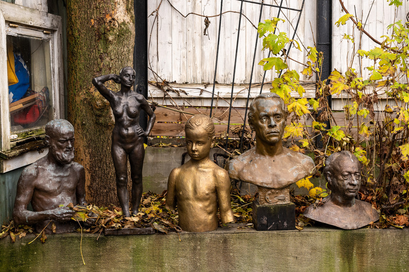 Utvidet bildebeskrivelse (for synshemmede):
Bildet viser en gruppe på fem realistiske skulpturer plassert utendørs langs en lav stein- eller betongmur. Skulpturene er utført i bronse og metall, med ulike overflater og patina. Fra venstre ses en eldre mann med skjegg, fremstilt i halvfigur, med hodet svakt bøyd. Ved siden av ham står en naken kvinneskikkelse i helfigur, med én arm bøyd bak hodet og en avslappet, selvsikker kroppsholdning. Midt i gruppen står en barnebyste i gyllen bronse, med kort hår og et alvorlig, konsentrert uttrykk. Til høyre for barnet står en mannsbyste med markerte ansiktstrekk og rolig blikk, montert på sokkel. Lengst til høyre ses en eldre mannsbyste med rynket ansikt og lukket, ettertenksom mimikk.

Bak skulpturene vokser en stor trestamme og visne klatreplanter med gule høstblader. Bakgrunnen består av en slitt, hvitmalt trevegg med synlige sprekker og aldring, noe som gir bildet en stemning av tid, historie og forgjengelighet. Skulpturene fremstår som studier av menneskekropp, alder og karakter, og samspillet mellom kunst og natur gir en rolig, poetisk atmosfære.

SEO-optimalisert bildebeskrivelse :
Utendørs gruppe av figurative skulpturer i bronse og metall, bestående av byster og helfigur av kvinne, menn og barn. Klassisk realistisk billedhuggerkunst plassert i ateliermiljø med høstløv, trær og værslitt trebygning i bakgrunnen. Skulpturene viser menneskekropp, portrettkunst og anatomiske studier i bronse.