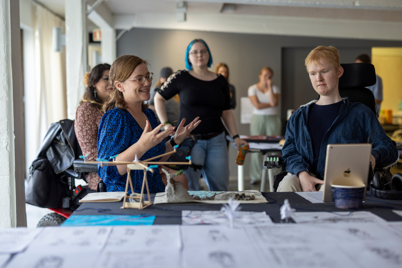 Flere mennesker i et rom ser mot et bord med steiner, modeller, tegninger og en iPad. En kvinne snakker til de andre personene i rommet og ser mot iPaden. To personer sitter i rullestol og flere står oppreist i bakgrunnen.