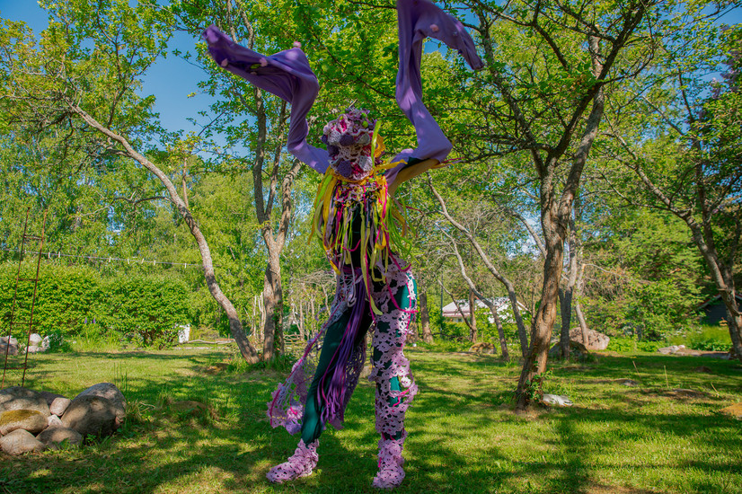 En stående skikkelse kledd i lilla kostyme løfter sleng-armene over hodet, ansiktet er dekket av en heklet blomst. Buksene har store lilla blonder over en smaragdgrønn thights.
Lange, gule, rosa og neongrønne tøyremser slenger fra en flettet krage, Figuren står midt i en irrgrønn vår-hage med trær.