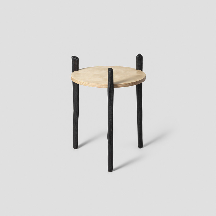 Rund bordplate i bjørk kryssfiner, trefarget med tre bein i ask, brennt, svart. Beina er uregelmessing og børstet, teksturen av treverket kommer tydelig frem.