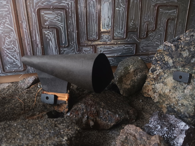 Bildet viser et tablau bestående av sand og små steiner, som i et landskap. En stein har en kobberspole viklet rundt seg, med en magnet og en kjegleformet megafonform festet mellom spolen og steinen. I bakgrunnen kan man se et metallrelieff som ligner et flytskjema.

