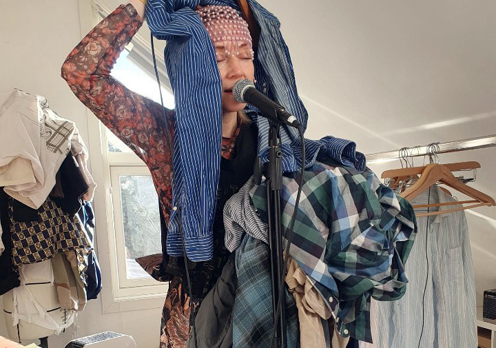 Et bilde av Turid Alida som synger i mikrofon med lukkede øyene. De holder en haug med klær på sin venstre arm og med sin høyre hånd holder de en stripete, blå skjorte som henger over deres hode. I bakgrunnen ser man en utstillingsdukke med flere lag med klær, et vindu og en klesstativ med en skjorte og flere tomme kleshenger.