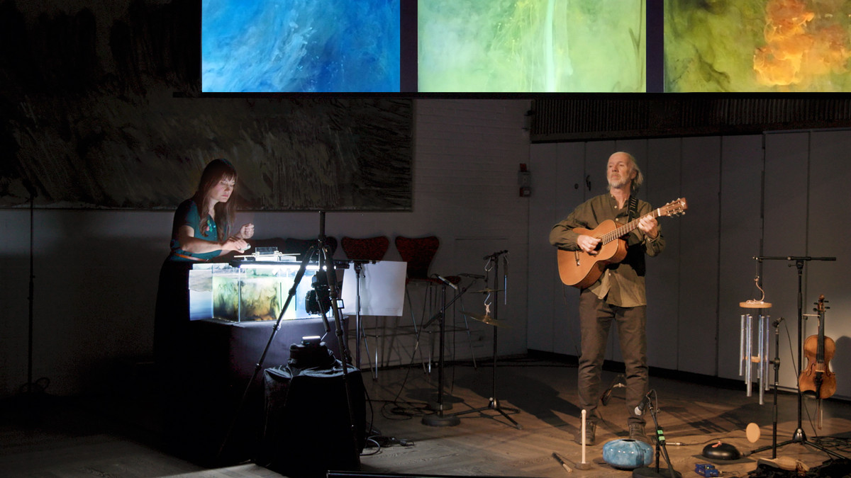 Til høyre en kvinne foran sterkt belyst akvarie fyllt med farget vann, til venstre en mann med guitar omringet av andre små musikkinstrumenter. I bakgrunnen tre projiserte lærreter med i blått, grønt og oransje.
