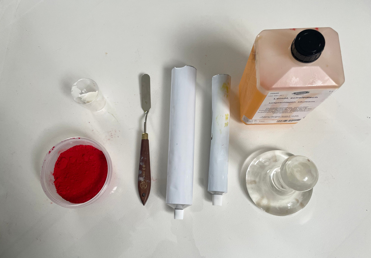 Rød pigment, en palettkniv, to tomme metalltuber, en flaske linolje og et glassinstrument på et hvitt bord