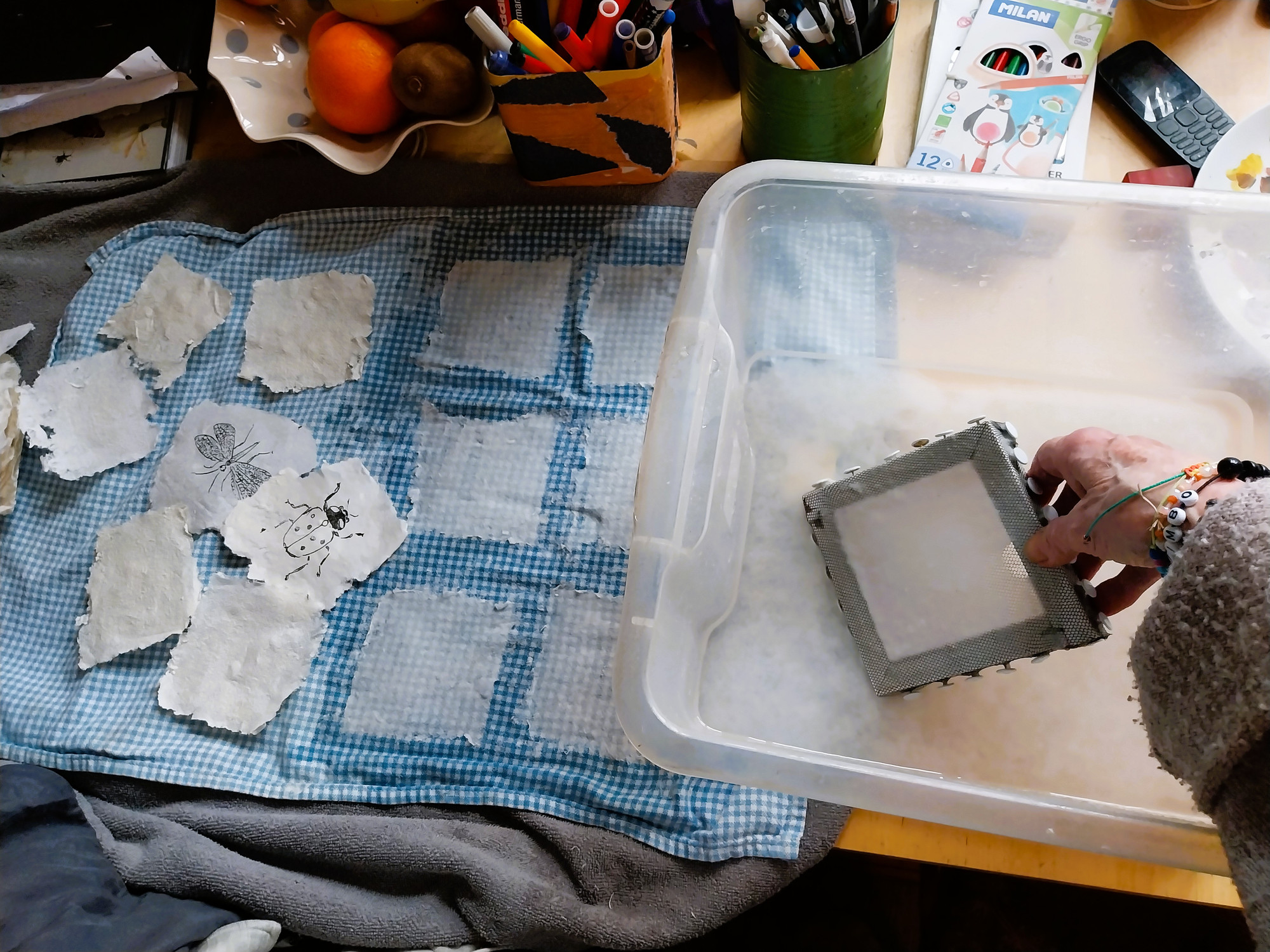 Prosessbilde av å lage papir på et bord fyllt med kunstmaterialer og andre små gjenstader.