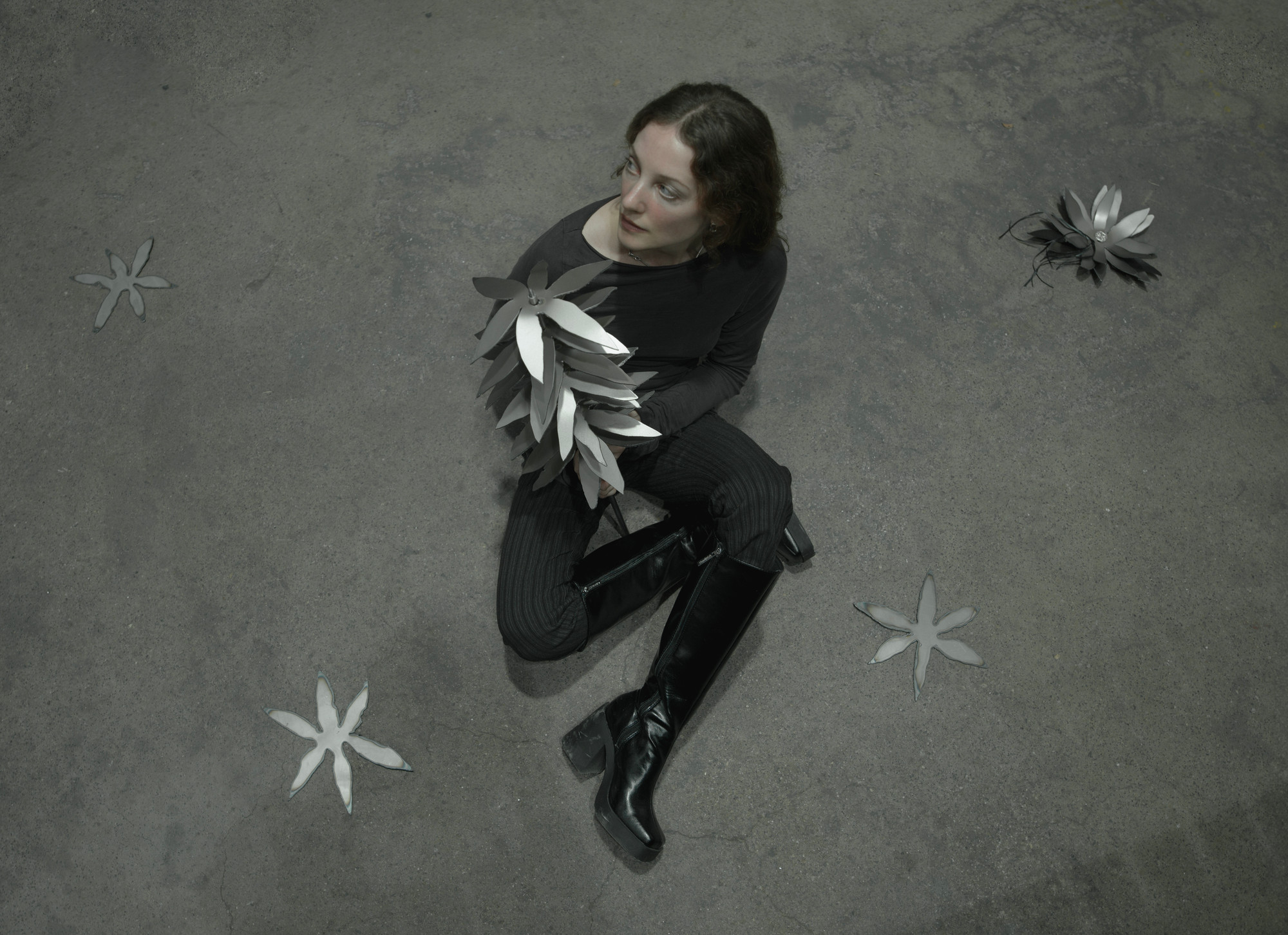 Kunstneren kledd i svart holder en blomsterlignende metallskulptur og sitter på et grått gulv. Rundt henne finnes flere deler som ligner skulpturen hun holder