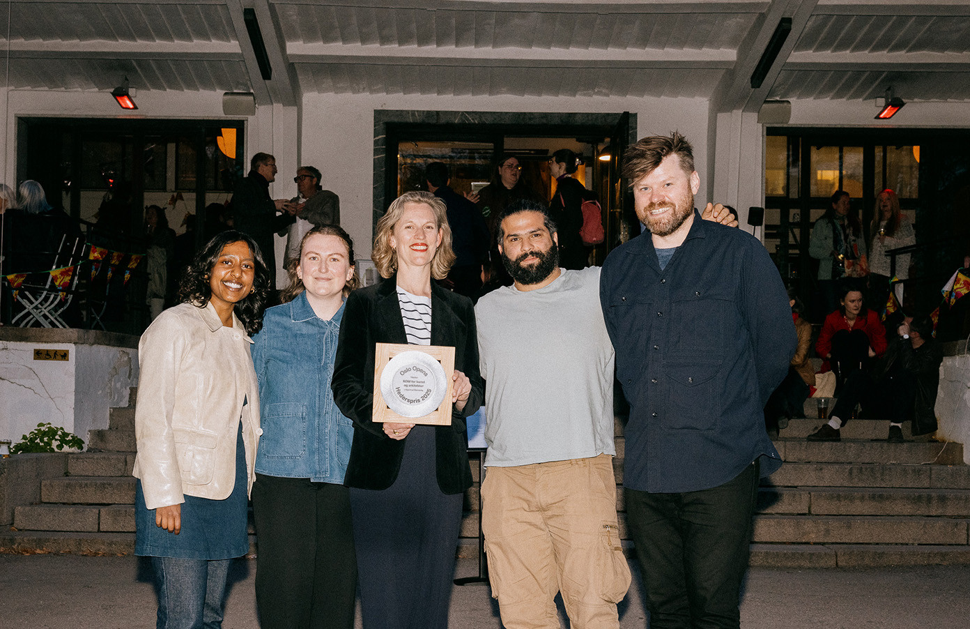 Fem personer smiler mot kamera og vider frem en pris-plakett. Bak dem foregår en fest. Det er kveld.