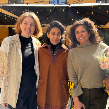 Tre kvinner smiler og poserer utendørs foran en gul bygning. Kvinnen til venstre har lyst, skulderlangt hår og er iført en lys, mønstret jakke over en svart genser og et langt, mørkt skjørt. Kvinnen i midten har mørkt, skulderlangt hår og er iført en brun tunika med broderier over en stripet genser. Kvinnen til høyre har mørkt, krøllete hår og holder en stor bukett med lyse roser innpakket i plast. Hun er iført en grå genser og mørke bukser. Bak dem er det en gul bygning med flere vinduer og svarte markiser med lyslenker under. Bakken er dekket av lyse steinheller.