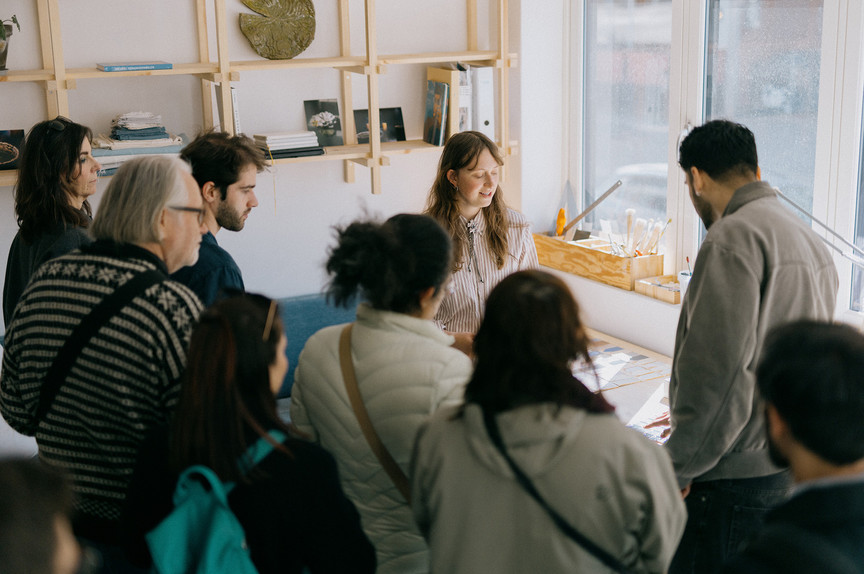 Kunstneren, en ung kvinne, omkranset av nysgjerrige besøkende. Rundt dem ser man materialer, verktøy og bøker. 