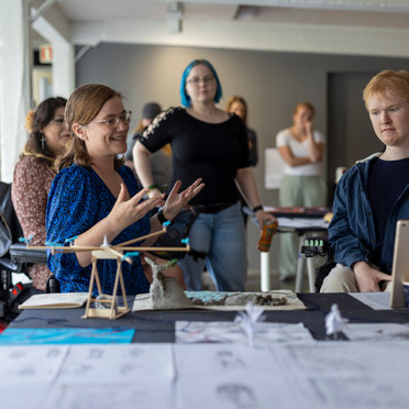 Flere mennesker i et rom ser mot et bord med steiner, modeller, tegninger og en iPad. En kvinne snakker til de andre personene i rommet og ser mot iPaden. To personer sitter i rullestol og flere står oppreist i bakgrunnen.