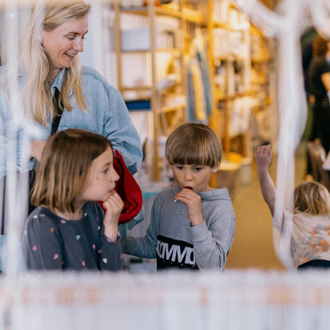 En mor og to barn kikker på en vev inne i et verksted.