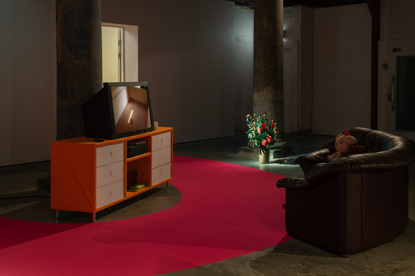 Installation view of Paradise Air: a brown leather sofa faces a small paper television on a paper table, surrounded by paper objects such as a remote, toilet roll and bottles, while images of a former love hotel architecture play on the screen.