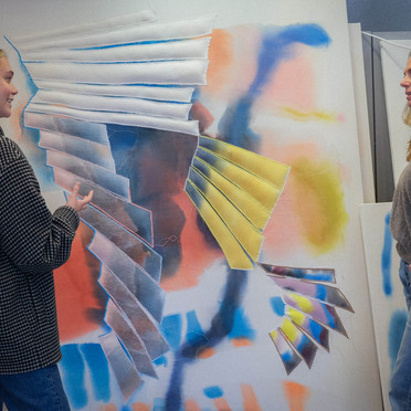 Marie Saxeide og kunstner Liv Tandrevold Eriksen snakker sammen foran et stort og fargerikt kunstverk som står lent opp mot en vegg.