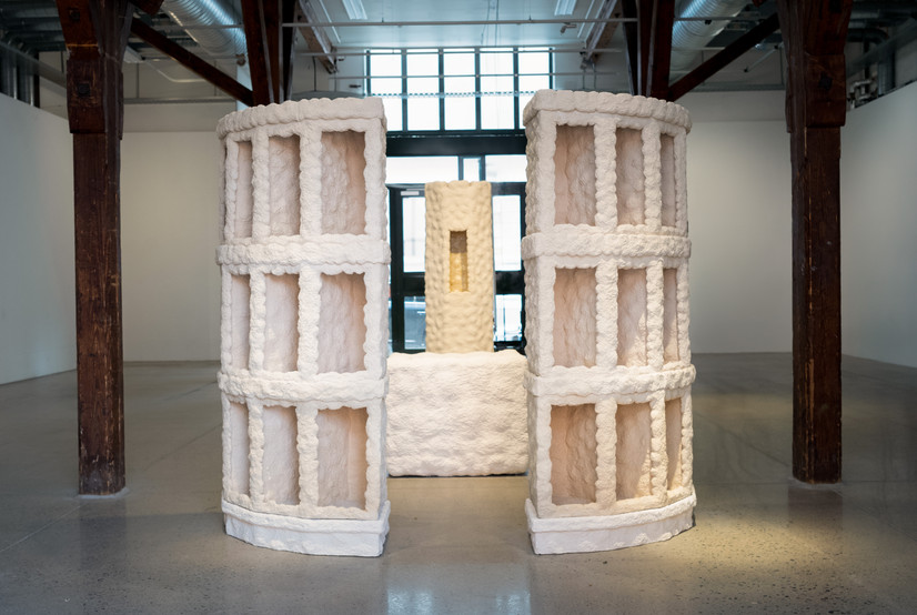 Overview of an installation consisting of four large ceramic sculptures. The sculptures have a rough surface and are unglazed. Each sculpture shares the same color, reminiscent of beige sand. The installation is arranged to guide you from one sculpture to the next, somewhat like a church aisle, with a highly symmetrical layout.

You first move through two identical sculptures that evoke the feeling of passing through a portal. Next, you encounter a rectangular basin that reaches hip height. This basin is filled with water. Finally, you arrive at the last sculpture, which resembles a tower in a castle. At the center of the tower is a chamber that extends inward. This chamber is glazed with a smooth, transparent glaze, resembling the water surface in the earlier basin-like sculpture. The entire installation is illuminated with warm light, evoking the ambiance of sunlight.