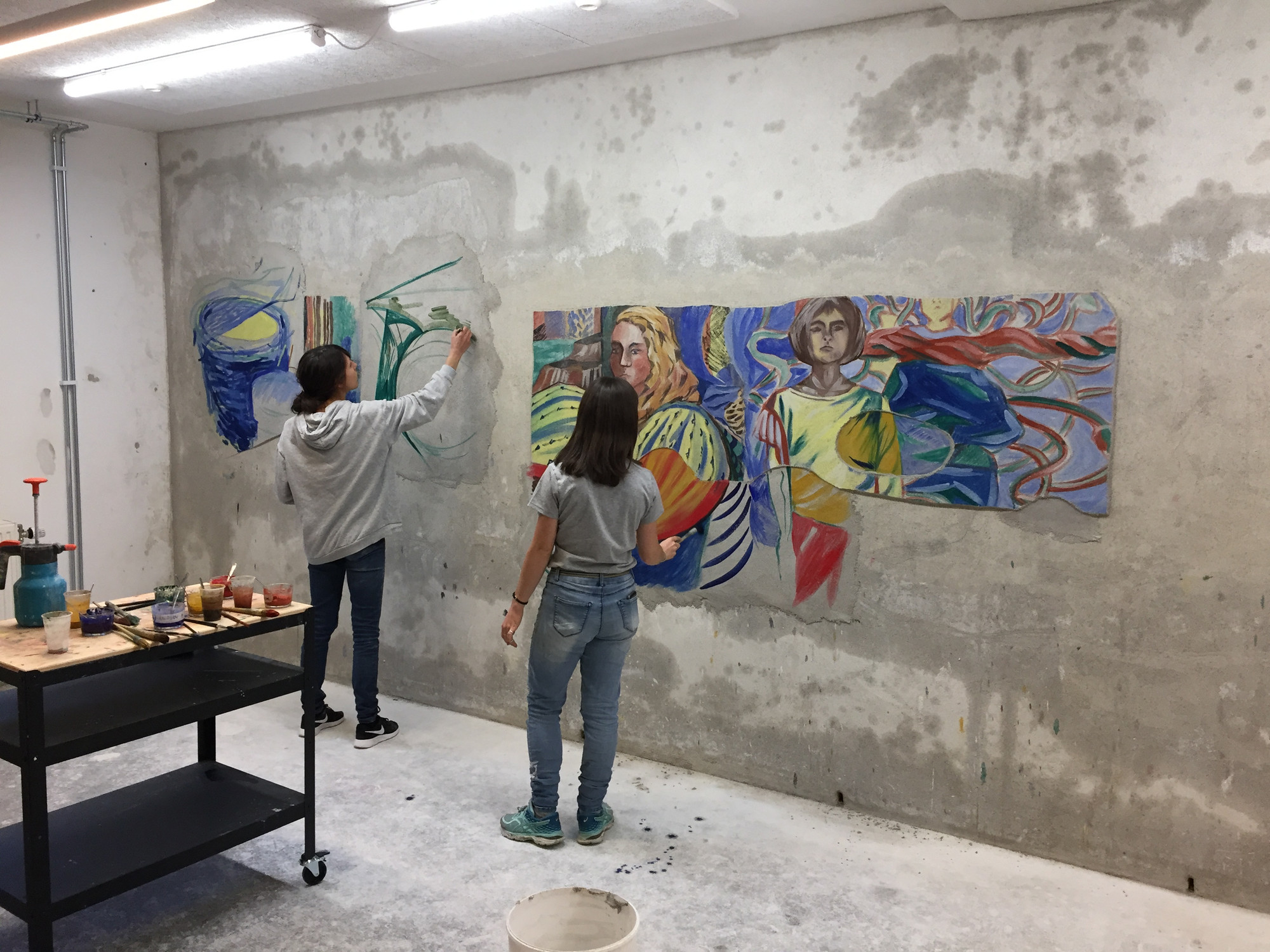 Ett grått rom hvor to personer- Mari Kolbeinson og Laura Gaiger- lager malerier rett på ene veggen. Bak de står det en tralle med kopper med maling og diverse maleutstyr.
