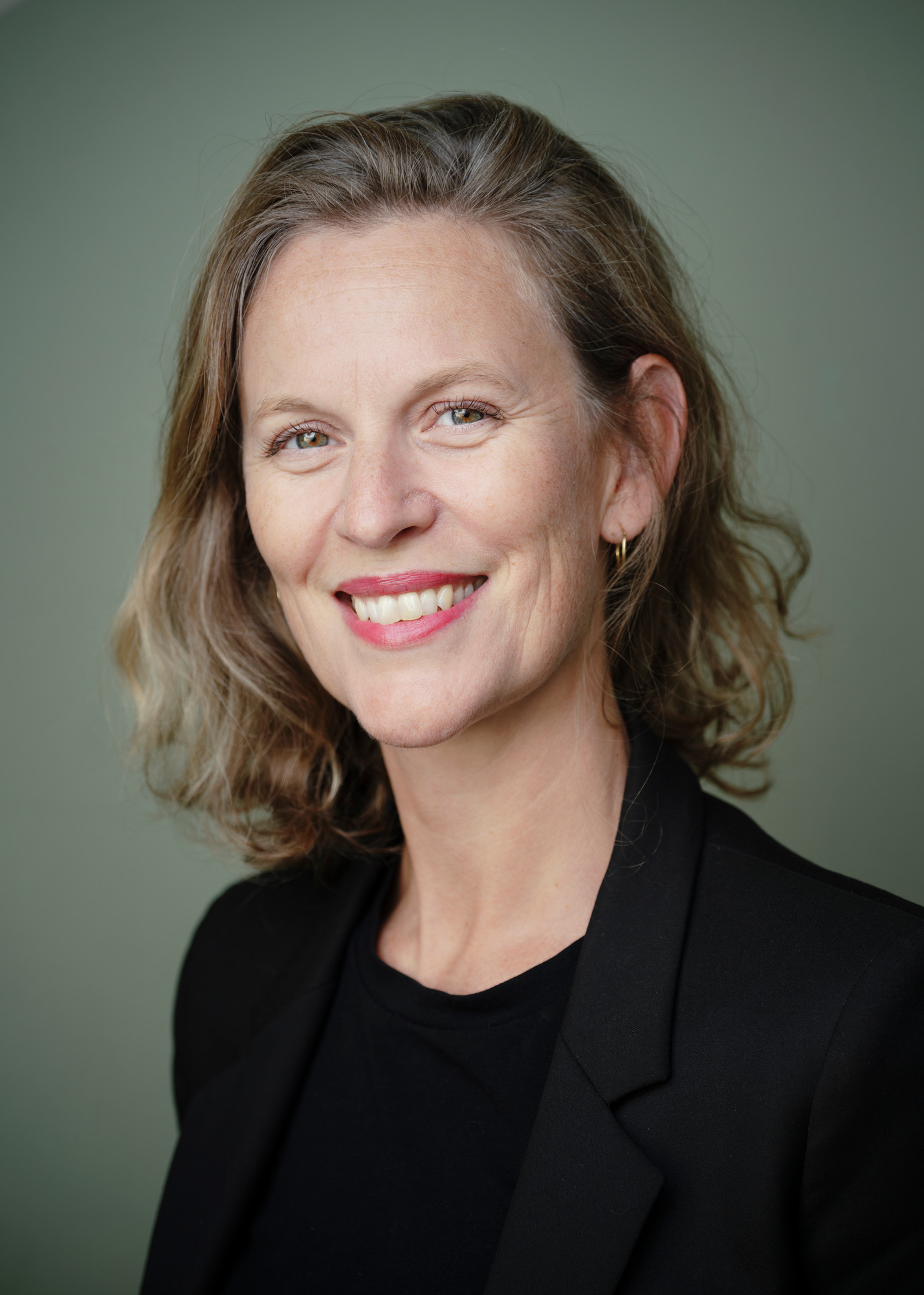 Portrettbilde av en kvinne med brunt hår i nakkelengde. Hun har på en sort skjorte med en sort dressjakke. Hun har også på rød leppestift og øreringer.