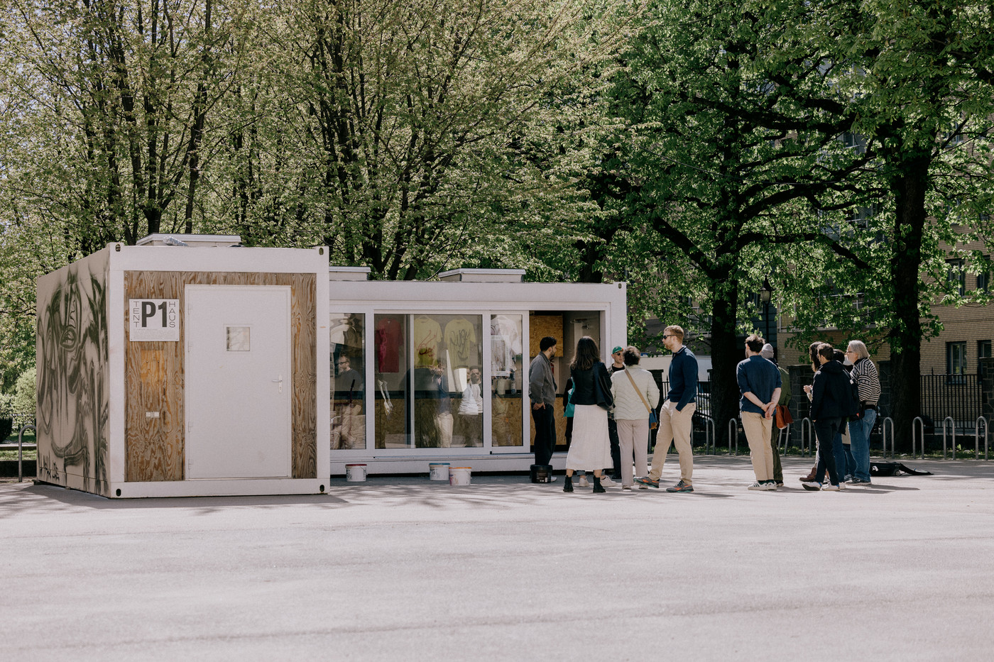 En gjeng med folk står og snakker utenfor et lite atelierbygg på en solig vårdag.
