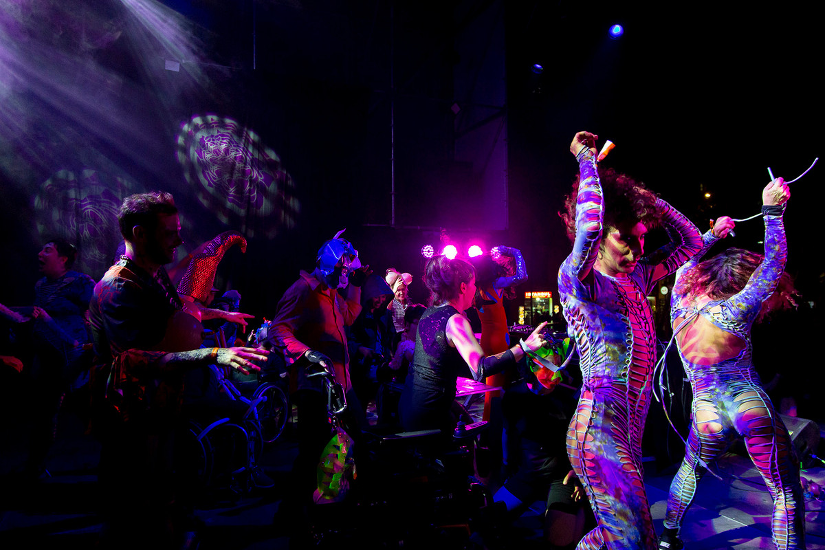 People is suggestive costumes, with and without wheelchairs, dancing under coloured lights.