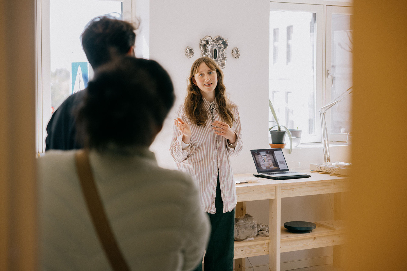 En ung kvinne tar imot to gjester i atelieret. I bakgrunnen en laptop og skulpturer.