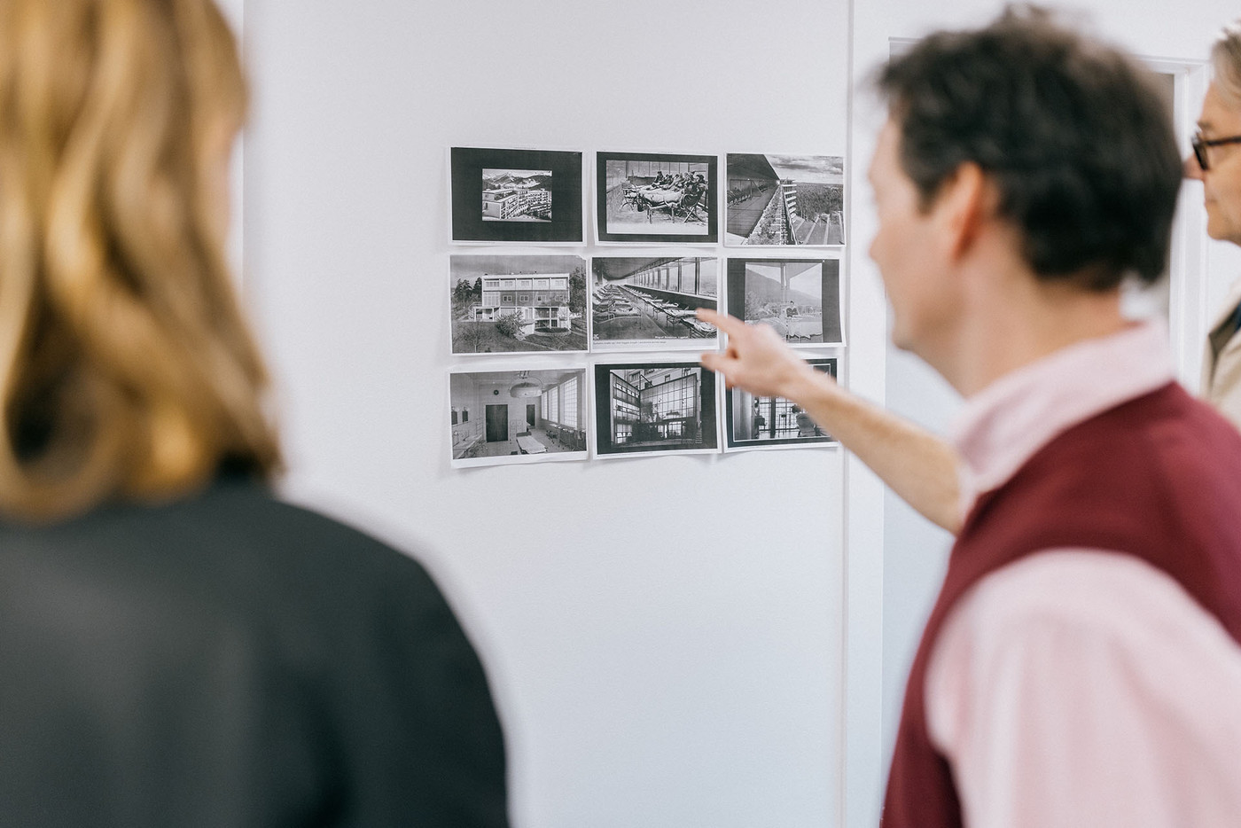 En mann med kort bustete hår viser fram svart/hvitt-fotografier av interiører og landskap til to besøkende.