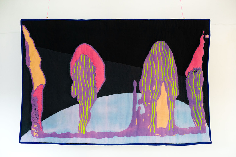 The work shows figures in pink, yellow, purple, orange and neon green, it can look poisonous. At the bottom is a half circle in light blue, and the background consists of black and gray that are divided diagonally from corner to corner. The work is hand-quilted.