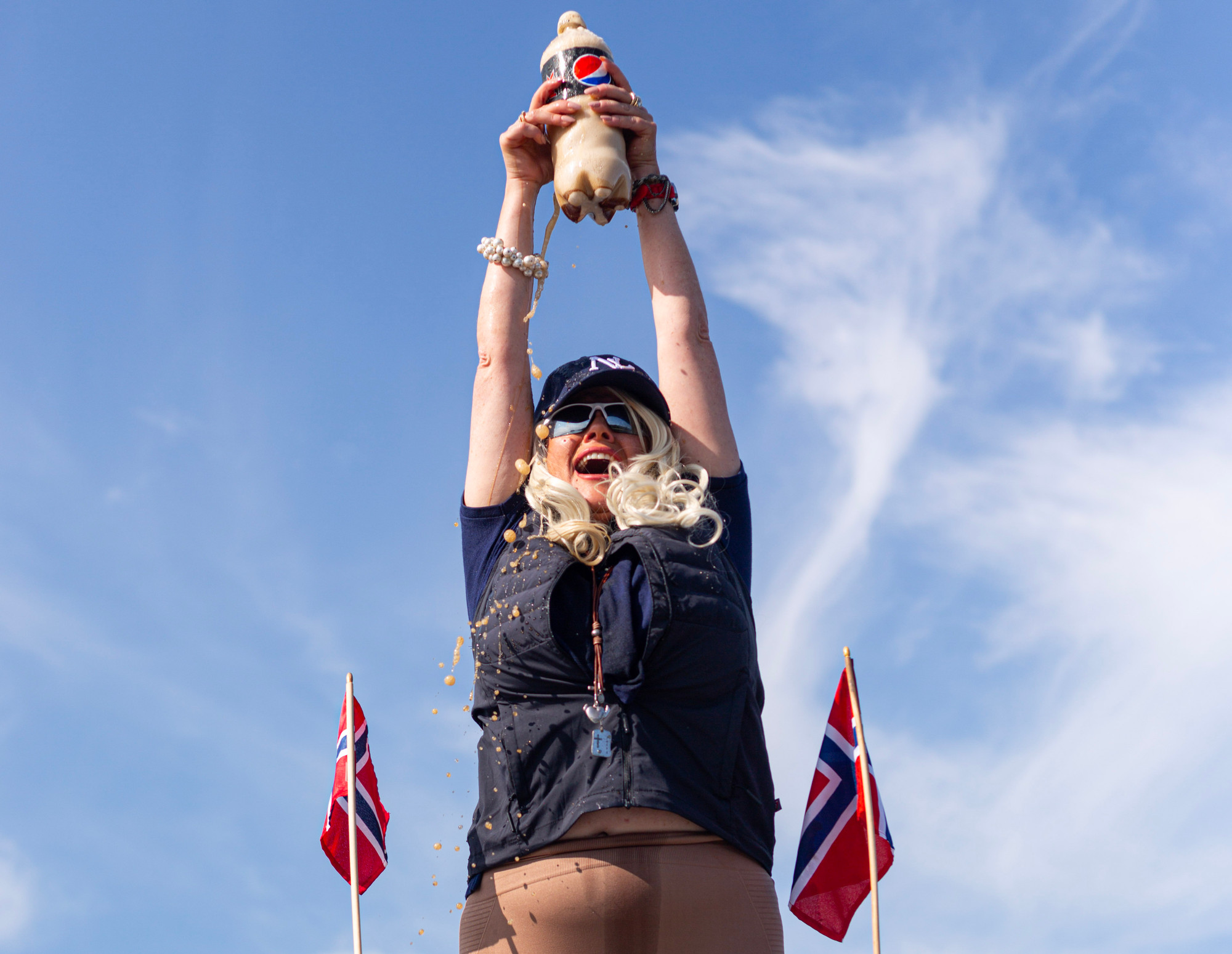En kvinne med bleket hår og solbriller holder opp en stor flaske med Pepsi Max mot en klarblå himmel med to norske flagg rundt seg