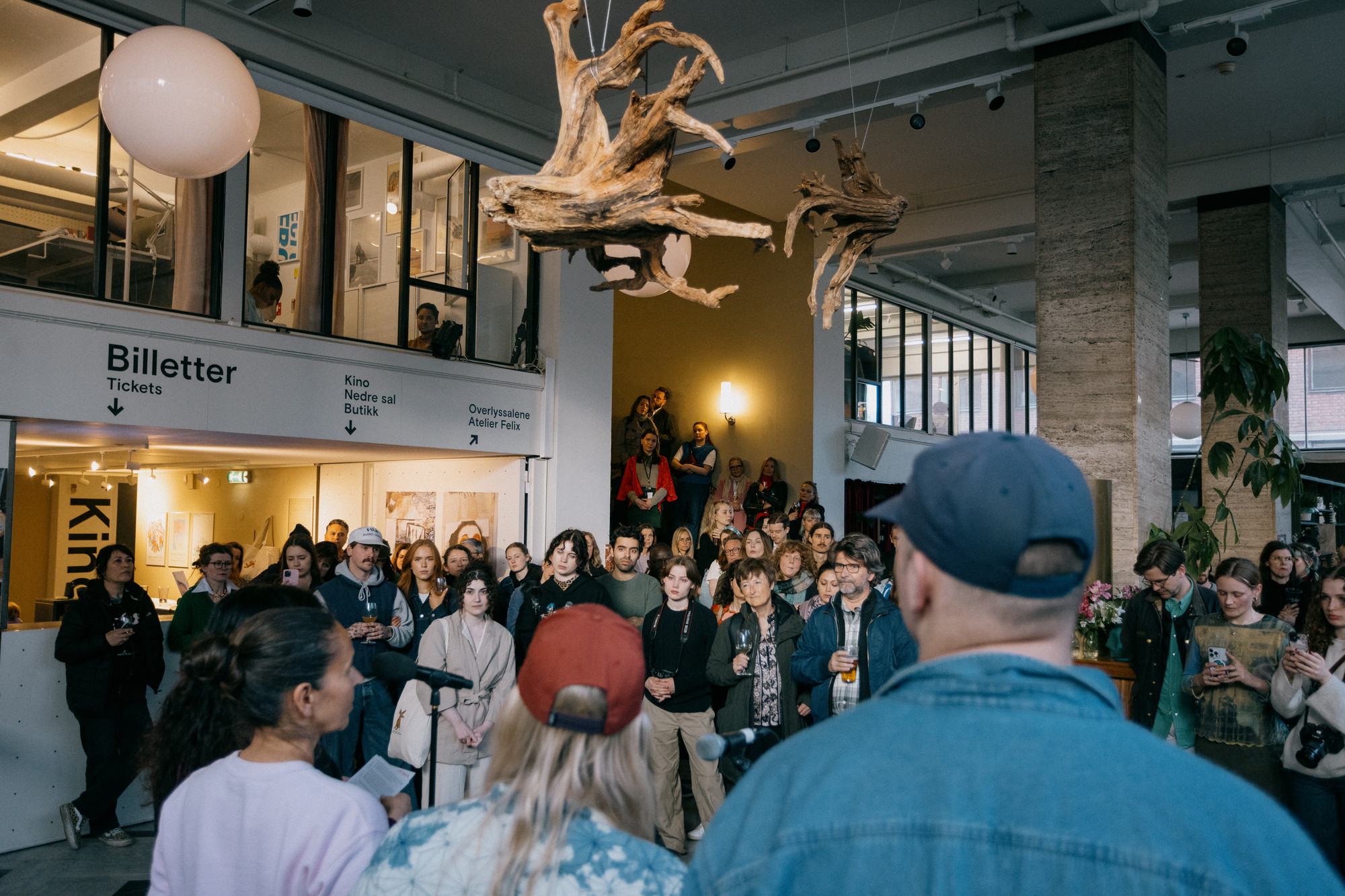 To kvinner og en mann står foran en folkemengde. Kunst henger fra taket. 