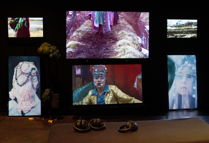 Et mørkt rom med to rader med tre flatskjermer.  Fire flatskjermer er vannrette mens de ytterste og nederste flatskjermene står på høykant på gulvet. Det to største TVene er i midten. På skjermene vises personer med masker forran asnsiktet, i bakgrunnen skimter en utendørs vår-lansdskap med snødekte Loffotfjell. Forran skjermene ser vi halvparten av en mørk tekstilkledd benk hvor det ligger tre høretelefoner. I mørket mellom noen av TVene skimeter vi masker laget av tekstil, farget fugeskum og perler, som blir brukt i videoene.