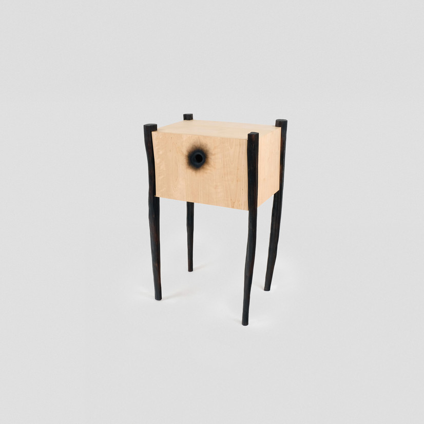 Møbel på fire bein. Kasse i bjørk kryssfiner, trefargen med skuff, gripehullet er brennt inn. Bein i kløyved heltre bjørk, brent svart, uregelmessig form.