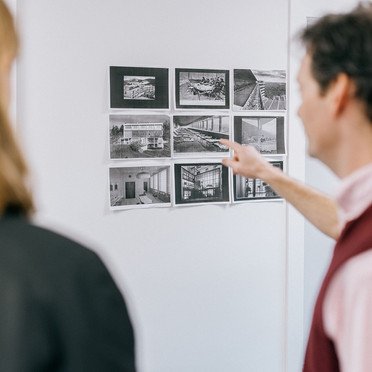En mann med kort bustete hår viser fram svart/hvitt-fotografier av interiører og landskap til to besøkende.