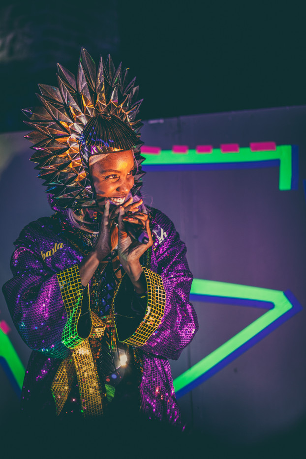 Black American woman stands smiling wearing a large shiny golden crown and a purple and gold sequined boxing  robe with a shiny gold corset underneath.