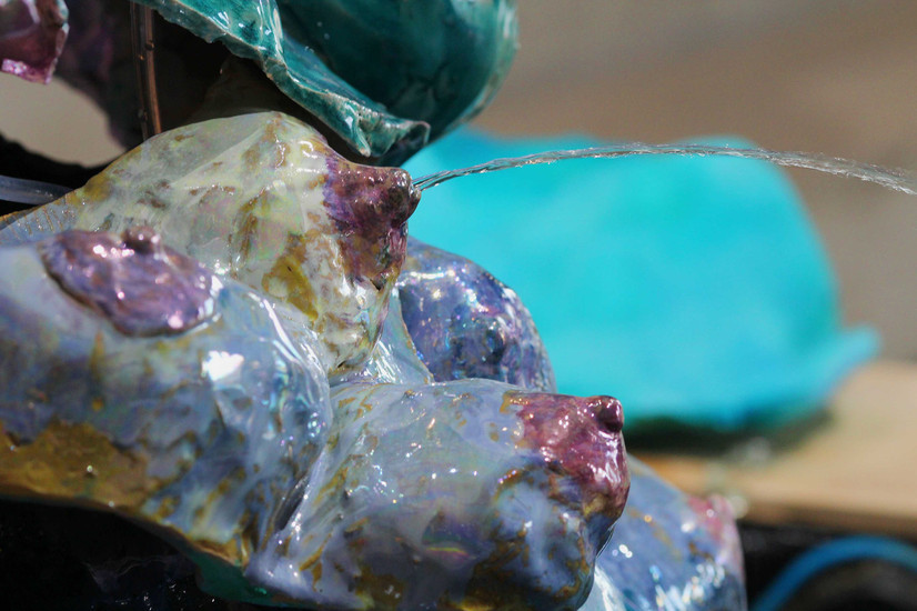 Nærbilde av en keramikk skulptur, vi ser fem bryster i gul eller lyseblå glinsende glasurer som ligner smeltende iskrem, som også har perlemors skimmer. Brystvortene er lilla eller mørkerosa, kunn to av dem er i fokus, de andre er blury. Ut av det øverste brystet til venstre, spruter det ut en buet vannstråle mot høyre side av bildet.